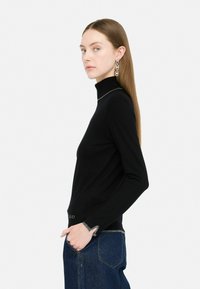 Zwarte turtleneck trui met lange mouwen, voorzien van zilverdraden in de manchetten en de zoom. Gecombineerd met donkerblauwe denim. Model staat zijwaarts.