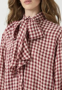 Gecheckte blouse met een rood-wit patroon, hoge kraag met een grote strikdetail en een lichte stoftextuur.