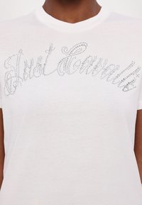 Bílá bavlněná tričko s jiskřící, kamínkovou výšivkou nápisu "Just Cavalli." Kulatý výstřih a krátké rukávy.