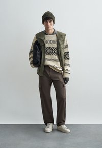 Kesztyűs krém színű pulóver fekete geometriai mintákkal, olajzöld mellény, barna cargo nadrág, szürke kesztyű és bézs sportcipő, zöld sapkával a tetején.