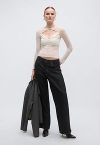 Λευκή μακρυμάνικη μπλούζα από δαντέλα με άνοιγμα, φορεμένη πάνω από ένα εκρού μπραλέτο. Σκούρο, φαρδύ τζιν παντελόνι και γκρι παλτό, συνδυασμένα με μαύρα μυτερά παπούτσια.