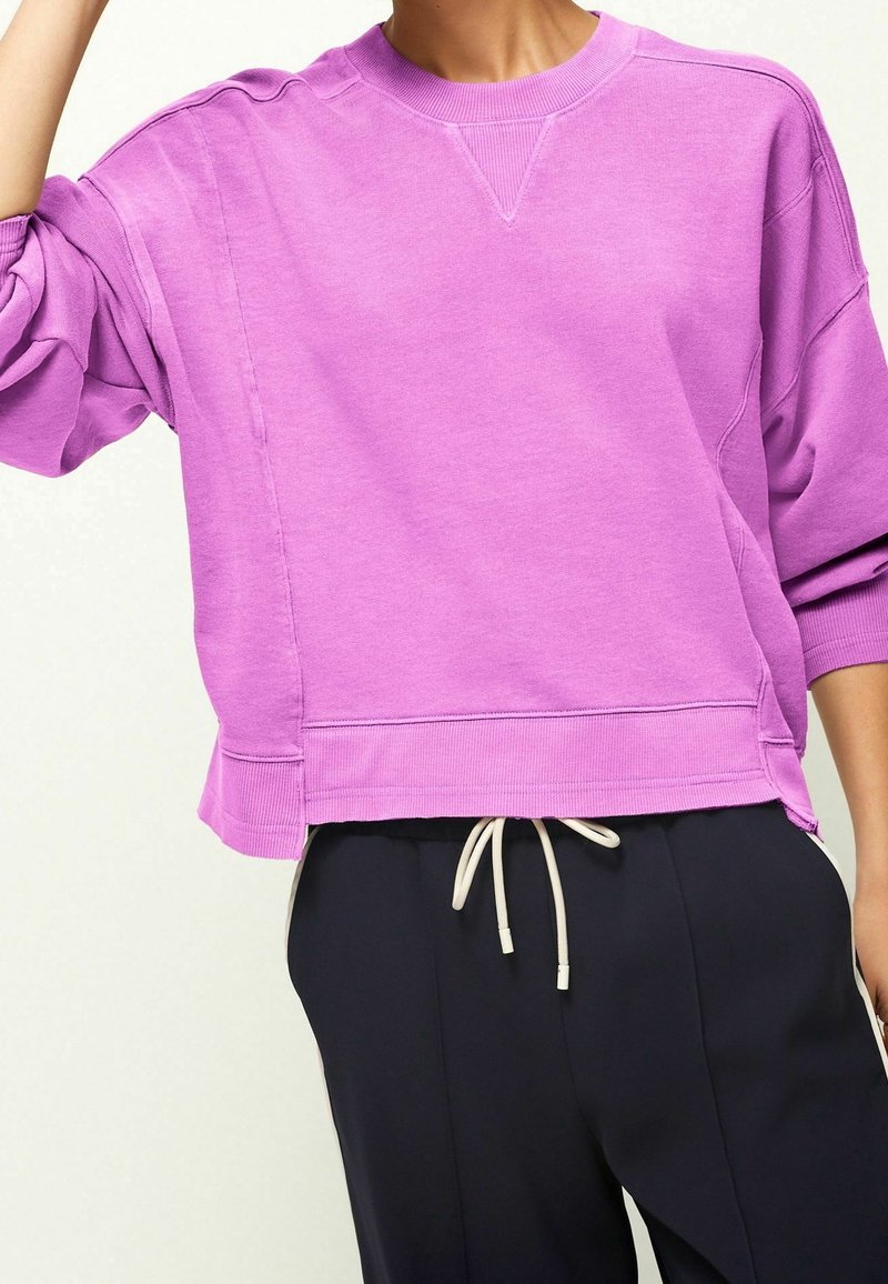 Personne portant un sweat-shirt ample lilas clair avec poignets et ourlet côtelés, associé à un pantalon noir avec cordon.