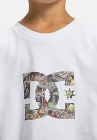 Biała bawełniana koszulka z kolorową grafiką przedstawiającą fantazyjne postacie i krajobrazy wewnątrz dużego nakładającego się logo DC.