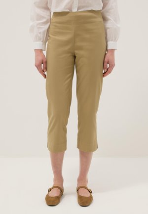 Mujer con pantalones beige recortados, blusa blanca de manga larga y zapatos planos Mary Jane marrones, de pie frente a un fondo liso.