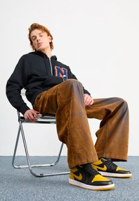 Svart zip-up hoodie med stor orange "N" logotyp, bruna vida byxor och svarta/gula Nike-sneakers, sittande på en kromstol mot en enkel bakgrund.