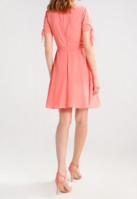 Korallklänning med korta ärmar knutna vid axlarna, en figursydd midja och en utsvängd kjol. Tyget är lätt ochsmidigt.