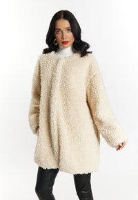 Palton oversized de culoare crem, realizat dintr-un material texturat și pufos, cu mâneci lungi și croială dreaptă, asortat cu pantaloni negri din piele.