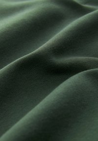 Dunkelgrüner Stoff mit glatter Textur und leichtem Stretch, der weiche, abgerundete Falten aufweist. Gleichmäßige Farbausbreitung mit minimal sichtbaren Mustern.