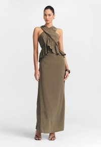 Maxime dress in oliivinvihreä, jossa on röyhelömäinen epäsymmetrinen bodice, yksi olkahihna ja sileä pinta. Yhdistetty mustiin hihnatossuihin.