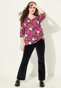 Lebendige lila Bluse mit floralen Mustern in Orange und Weiß, kombiniert mit schwarzen Cordhosen und schwarzen klobigen Schuhen.