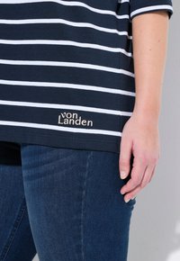 Maglietta a righe navy con strisce bianche, caratterizzata da un logo ricamato "von Landen" in beige all'orlo, abbinata a jeans blu scuro.