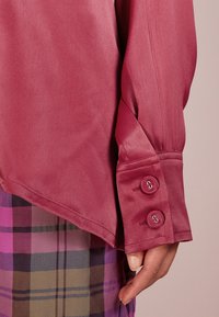 Krátký rukáv vínové blůze s hladkou texturou, vybavené manžetami na dva knoflíky. Pod tím se nachází vzorovaná látka s kostkovaným vzorem v odstínech fialové a hnědé.