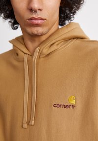 Brown hoodie made of textured fabric, featuring a front kangaroo pocket, drawstring closure, and embroidered Carhartt logo in yellow and burgundy.