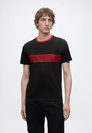 Joven con una camiseta negra con cuello rojo y rayas horizontales rojas en el pecho, de pie frente a un fondo blanco.