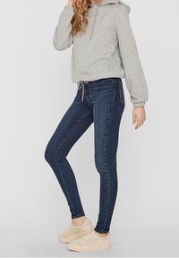 Sudadera con capucha gris de corte relajado, combinada con jeans ajustados azul oscuro y zapatillas color beige claro. El conjunto presenta un estilo urbano casual.
