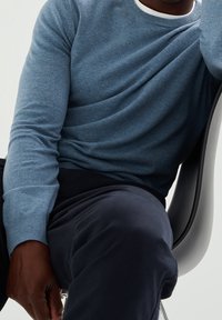 Blue knit sweater with a round neckline and long sleeves, paired with dark trousers, sits on a gray chair in a minimalist setting.