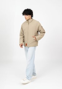 Jasnobeżowa lekka kurtka z wysokim kołnierzem, zamek błyskawiczny z przodu, kieszeniami po bokach oraz teksturowanym znaczkiem u dołu. Nosi ją na jasnoniebieskich dżinsach.