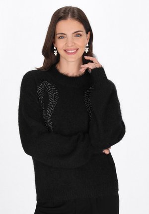 Smilende kvinde med langt brunt hår iført sort sweater med perleblad-design og hvid blomsterformet øreringe mod hvid baggrund.