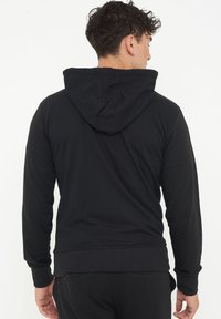 Felpa nera con cappuccio regolabile, maniche lunghe e polsini a costine. Il tessuto appare morbido con una texture liscia. Presenta una tasca a marsupio.