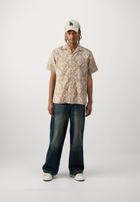 Camicetta beige a maniche corte con stampa floreale bianca, abbinata a jeans a gamba larga blu scuro e sneakers bianche, completata da un berretto crema.