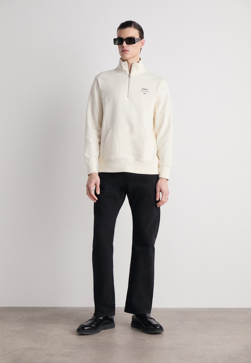 Felpa a zip corta color crema chiara con tasca frontale e logo, abbinata a pantaloni neri a gamba dritta e scarpe nere lucide.