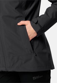 Giacca impermeabile nera con una superficie liscia, dotata di tasca laterale e polsini regolabili con Velcro, marchiata "Jack Wolfskin."