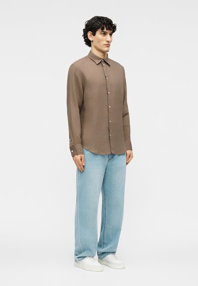 Άνδρας στέκεται φορώντας καφέ πουκάμισο με κουμπιά, ανοιχτό μπλε τζιν παντελόνι φαρδιάς γραμμής και λευκά αθλητικά παπούτσια μπροστά σε απλό ανοιχτό φόντο.