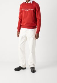 Czerwona bluza z kapturem z białym logo "TOMMY HILFIGER EST. 1985", w połączeniu z białymi spodniami i czarnymi butami typu slip-on. Swobodny, luźny krój.