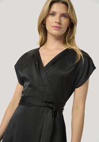 Musta satiini kietaisumekko, jossa on lyhyet hihat, V-pääntie ja solmittava vyötärö. Sujuva tekstuuri ja sulavalinjainen malli korostavat sen eleganttia ilmettä.