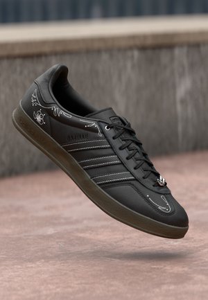 Baskets noires Adidas avec des motifs de coutures blanches, des lacets noirs et une semelle marron suspendues au-dessus d'une surface en béton rosâtre.