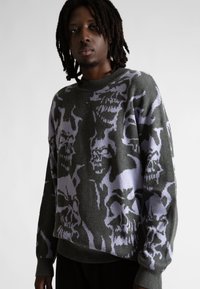 Mężczyzna z dredami, ubrany w szaro-lawendowy sweter w dużą abstrakcyjną czaszkę, stoi na jednolitym jasnym tle.