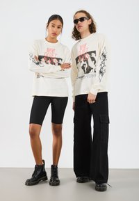 Cream long-sleeve shirts with graphic prints, paired with black biker shorts and cargo pants. Both wear black combat boots and sunglasses.