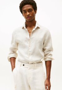 Camisa de lino a rayas de color beige claro con cuello tipo button-down y mangas arremangadas, combinada con pantalones de color crema que tienen un botón negro.