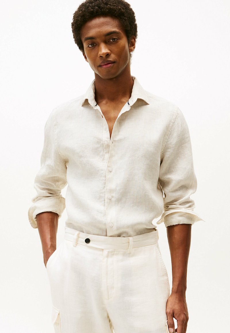 Camisa de lino a rayas de color beige claro con cuello tipo button-down y mangas arremangadas, combinada con pantalones de color crema que tienen un botón negro.