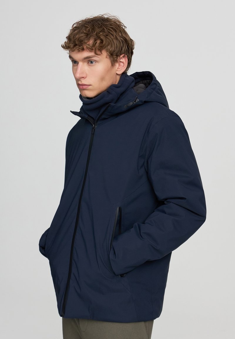 Giacca imbottita blu navy con cappuccio e zip frontale, dotata di tasche laterali e una texture liscia, progettata per mantenere il calore e resistere alle intemperie.