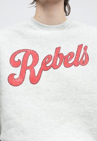 Pelēks džemperis ar lielu, nodilušu sarkanu uzrakstu "Rebels" kursīvā fontā priekšpusē. Mīksta tekstūra ar ribotu apkakli.