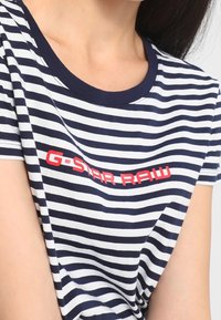 Camiseta de rayas azul marino y blanca con escote redondo, que presenta un audaz logo rojo "G-Star RAW" en el pecho. Material de algodón suave.
