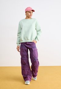Sudadera verde menta, pantalones cargo morados con múltiples bolsillos y una gorra rosa. Las zapatillas presentan una combinación de colores azul claro y gris.