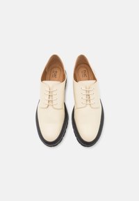 Scarpe in ecopelle beige con punta arrotondata, design con lacci e suola in gomma nera a contrasto. Finitura liscia e stile minimalista.