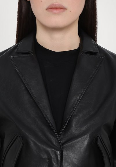 Black leather blazer with notched lapels, tailored design, and visible stitching. Worn over a black crew-neck shirt, showcasing a sleek texture.