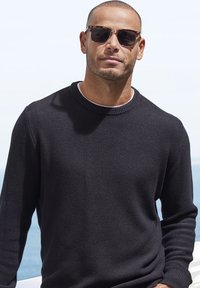Schwarzer Strickpullover mit strukturiertem Muster und rundem Halsausschnitt, kombiniert mit einer Schildpatt-Sonnenbrille, vor einem hellen Außenhintergrund.