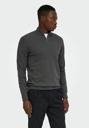 Young man wearing a dark gray half-zip sweater and black pants, standing and looking to his right against a neutral background.