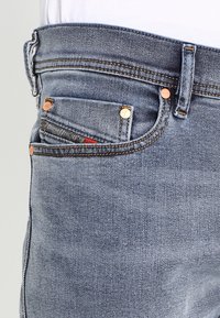 Jeans de mezclilla azul con corte ajustado, detalles en remaches de cobre y un pequeño bolsillo delantero con marca. Tejido texturizado con costuras visibles.