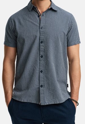 Mand iført kortærmet, navyblå og hvide lodrette stribede skjorte med knapper, med hænderne i lommerne på mørke bukser.