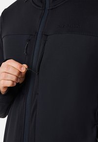 Zwarte jas met een rits aan de voorkant, voorzien van een hoge kraag, gladde textuur en een logo op de borst. Verstelbare trekkoord aan de onderkant.