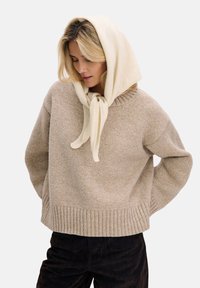 Beżowy sweter z dzianiny z prążkowanym wykończeniem u do�łu i luźnym krojem, zestawiony z jasnym kremowym szalikiem związanym na szyi. Miękka faktura, casualowy design.