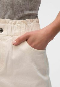 Pantalones color crema con una cinturilla elástica fruncida, un bolsillo lateral y una textura suave. Parte de una blusa gris con patrón es visible.