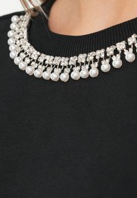 Prenda negra con un collar que presenta filas alternas de perlas y rhinestones claros, sobre una textura suave.