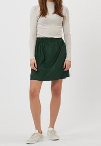 Jupe en nylon vert avec une taille élastique, un ourlet plat et une texture lisse, associée à un haut léger à manches longues et des baskets.