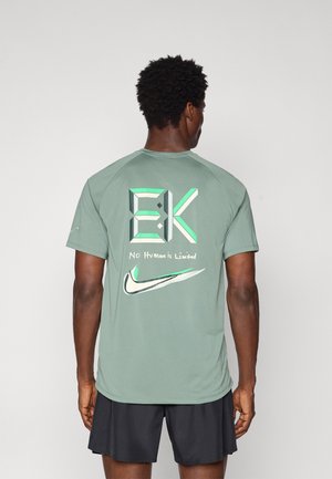 Zaļš sportisks krekls ar īsām piedurknēm, uz kura aizmugures ir liela grafika ar tekstu un Nike logo kontrastējošās krāsās.